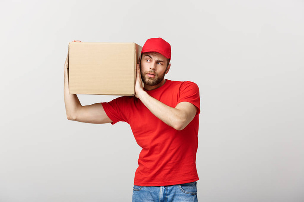 Delivery Concept - Portrait of Curious caucasian handsome delivery man listen inside a box package. Isolated on Grey studio Background. Copy Space. - Foto, Imagen