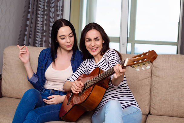 Cheerful beautiful girl friends playing guitar sitting on the couch. - Фото, зображення