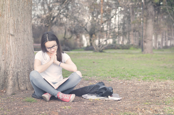 Beautiful young school or college girl with long hair, eyeglasses and black leather jacket sitting on the ground in the park reading the books and study for exam, education concept - Fotoğraf, Görsel