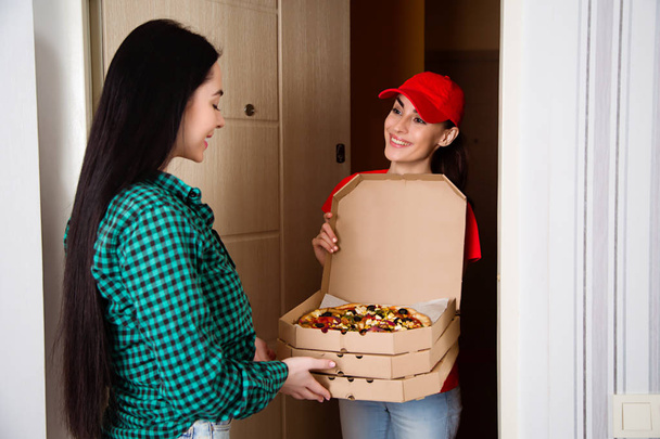 Young smiling girl courier in red uniforms delivered three boxes of pizza to the customer in her apartment or house. - Photo, image