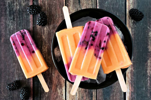 Kom van mango blackberry yoghurt ijslolly, bovenaanzicht op een donkere rustieke houten achtergrond - Foto, afbeelding