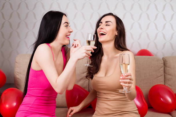 Panieński. Dwie piękne dziewczyny wesoły w jasne suknie wieczorowe pić szampana i reszta na imprezie, siedząc na kanapie. - Zdjęcie, obraz
