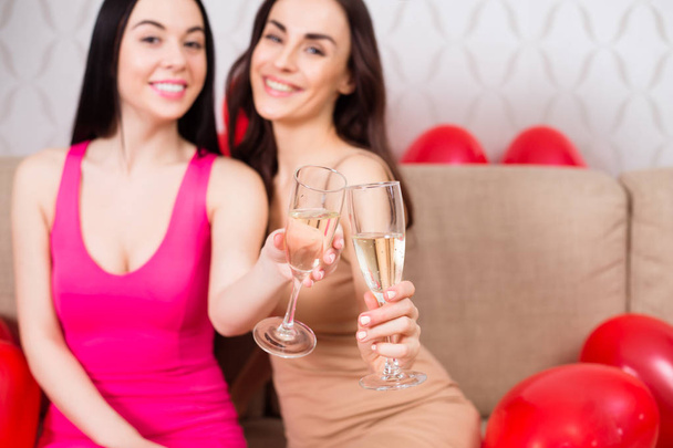 Κότα κόμμα. Δύο όμορφα κορίτσια εύθυμο σε φωτεινά φορέματα βραδιού πίνουν σαμπάνια και το υπόλοιπο σε ένα πάρτι που κάθεται στον καναπέ. - Φωτογραφία, εικόνα