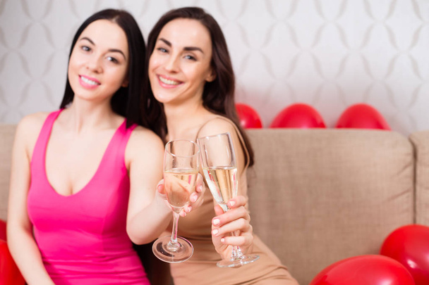 Куриная вечеринка. Две красивые веселые девушки в ярких вечерних платьях пьют шампанское и отдыхают на вечеринке, сидя на диване.
. - Фото, изображение