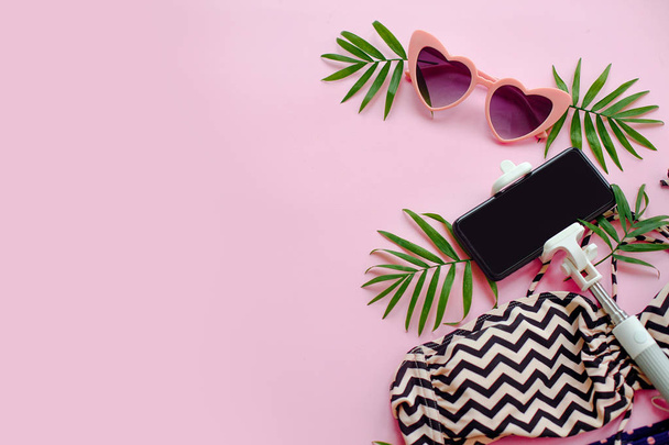 κομψό ροζ γυαλιά ηλίου, μοντέρνα μαγιό, τηλέφωνο selfie stick και φύλλα πράσινα φοίνικα σε ροζ φόντο, επίπεδη lay. χώρο για το κείμενο. καλοκαιρινές διακοπές έννοια. χρόνο να ταξιδέψει έννοια - Φωτογραφία, εικόνα