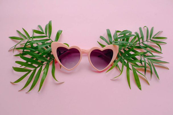 стильные розовые очки в форме сердца и зеленые пальмовые листья на розовом фоне, плоский лежал
.  - Фото, изображение