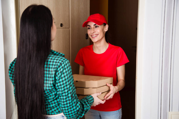 Young smiling girl courier in red uniforms delivered three boxes of pizza to the customer in her apartment or house. - Foto, imagen