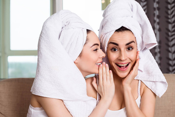 Beautiful smiling young  friends in white towels on their heads after a spa gossip or discuss something sitting on the couch. The girls are resting. - Fotoğraf, Görsel