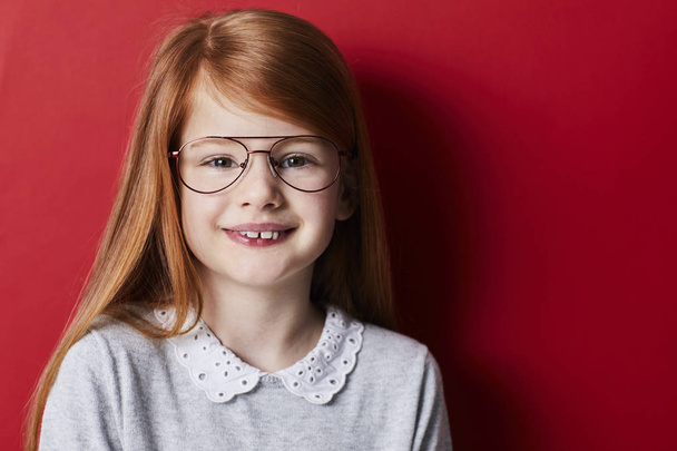 Portrait of redheaded girl in glasses smiling at camera on red background - Valokuva, kuva