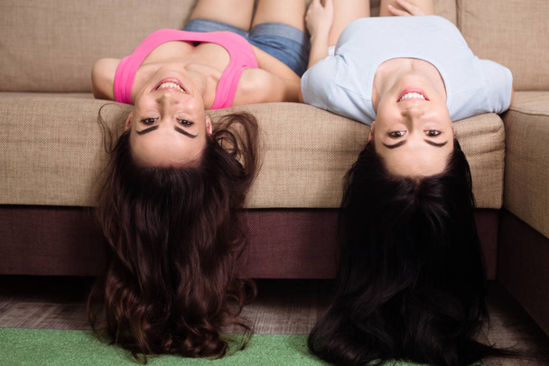Two funny and happy girl friends or sisters are lying on the couch upside down and having fun - Fotografie, Obrázek