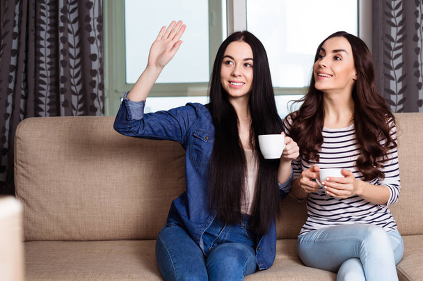 Two smiling beautiful women drink coffee or tea at home  and discuss the news - Фото, изображение