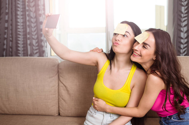  Beautiful cheerful girlfriends in casual clothes take a photo on the phone while sitting on the sofa at home. - Foto, Bild