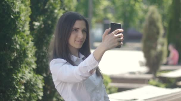 молода жінка фотографує себе на смартфоні вдень на відкритому повітрі. Дівчина робить селфі після обіду в міському парку
 - Кадри, відео