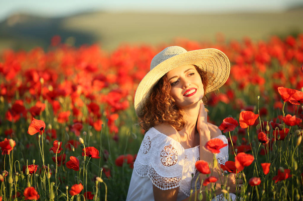 young beautiful woman posing on poppy field wearing white dress and straw hat  - Fotó, kép