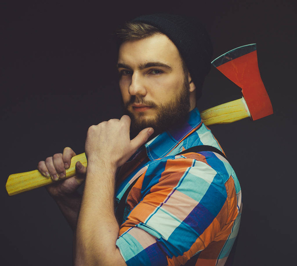 Serious lumberjack. Confident young bearded man holding a big axe and looking at camera while standing against black background. - Photo, Image