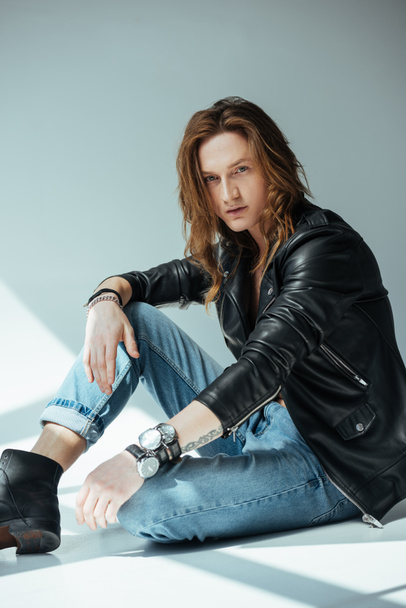 handsome man with long hair posing in jeans and black leather jacket, on grey - Fotoğraf, Görsel