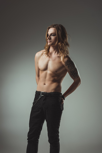 shirtless man with long hair and tattoo posing in black jeans, isolated on grey - Zdjęcie, obraz