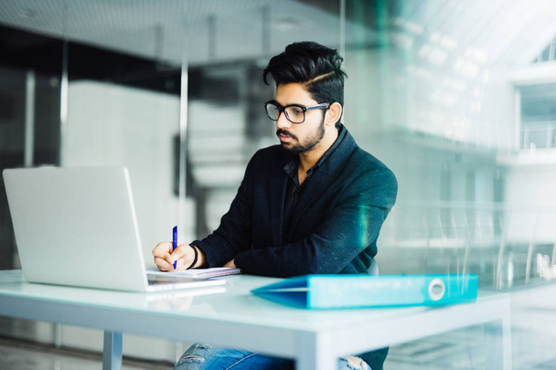 Веселый молодой индийский бизнесмен пишет в блокноте на столе в офисе
 - Фото, изображение