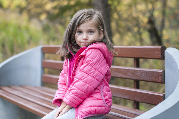 Little pretty girl in a pink coat sitting on a wood bench at the park in autumn. Emotional portrait. Childhood concept. Caucasian. Child with brunette long hair. Leisure, relaxation, lifestyle - Φωτογραφία, εικόνα