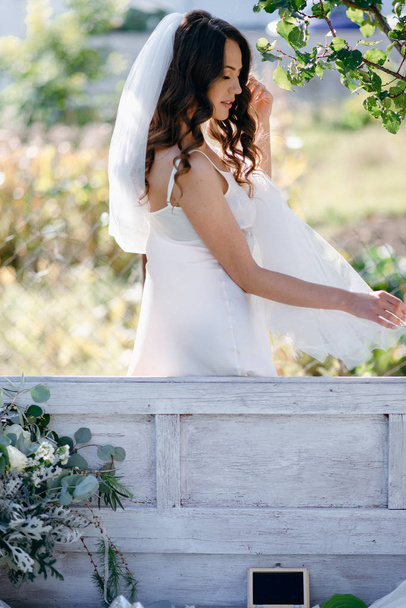 Портрет невесты с волнистыми волосами и вуалью, стоящей на скамейке в саду
 - Фото, изображение