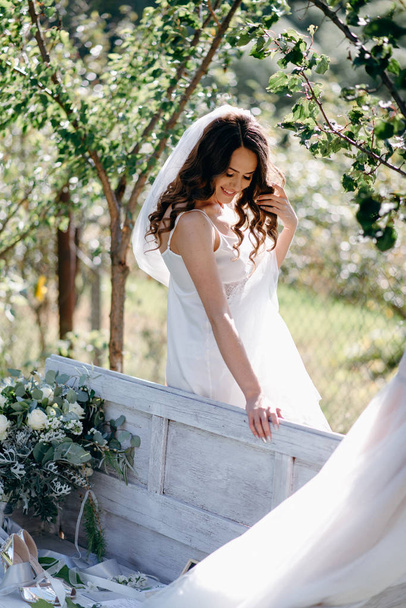 Πορτρέτο της νύφης με κυματιστά μαλλιά και το πέπλο στέκεται στο πάγκο στον κήπο - Φωτογραφία, εικόνα