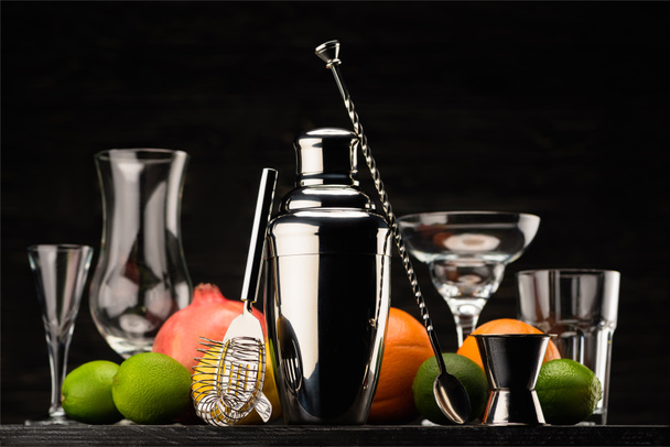 shining shaker for preparing alcohol drink, empty glasses and ripe fruits on table isolated on black - Photo, Image
