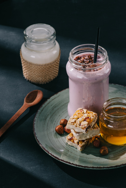 closeup view of berry smoothie with drinking straw, slices of energy bar, jam and walnuts on plate, spoon and bottle of yogurt on table  - Photo, Image