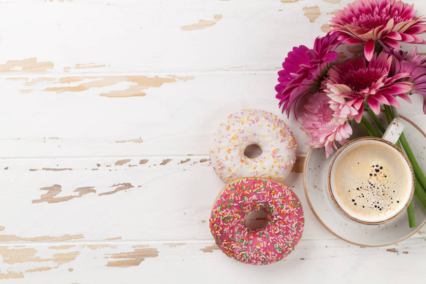 Taza de café, rosquillas y flores de gerberas en una mesa de madera blanca. Vista superior con espacio para su texto
 - Foto, Imagen