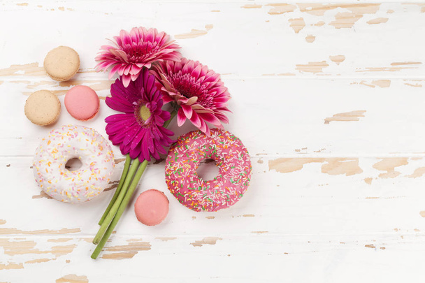 Donuts and gerbera flowers bouquet on white wooden table. Top view with notepad for your text - Φωτογραφία, εικόνα