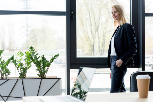 beautiful smiling blonde businesswoman standing with hand in pocket and looking away in office - Φωτογραφία, εικόνα