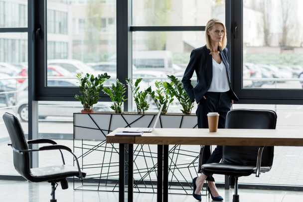 confident businesswoman standing with hands in pockets and looking away in office - Фото, зображення