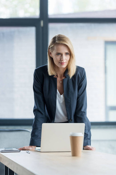 attractive blonde businesswoman leaning at table with laptop in office - Foto, Imagem