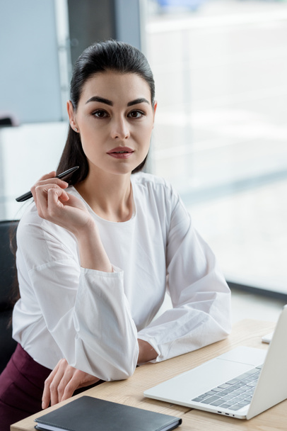 beautiful young businesswoman holding pen and looking at camera in office - Foto, afbeelding