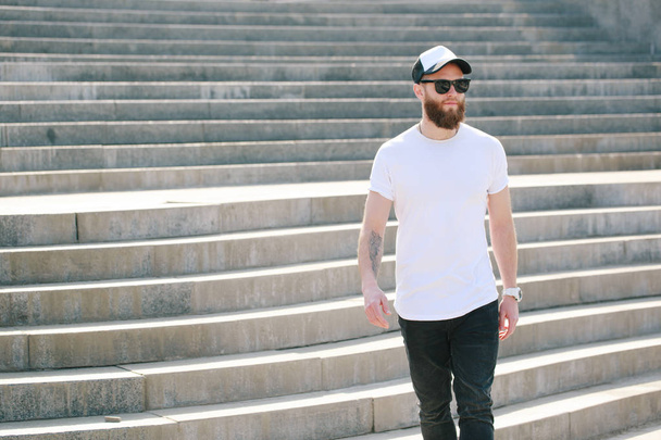 Hipster handsome male model with beard wearing white blank t-shirt and a baseball cap with space for your logo or design in casual urban style - 写真・画像
