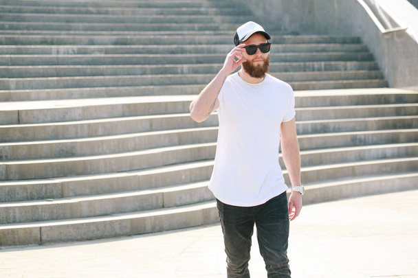 Hipster handsome male model with beard wearing white blank t-shirt and a baseball cap with space for your logo or design in casual urban style - Foto, Imagen