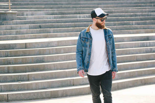 Hipster handsome male model with beard wearing white blank t-shirt and a baseball cap with space for your logo or design in casual urban style - Fotó, kép