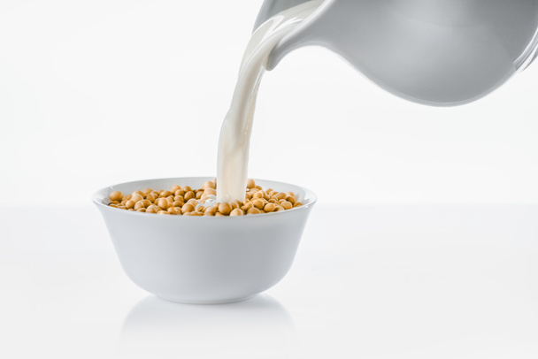 Pouring milk from jug in bowl with soybeans on white background - Zdjęcie, obraz