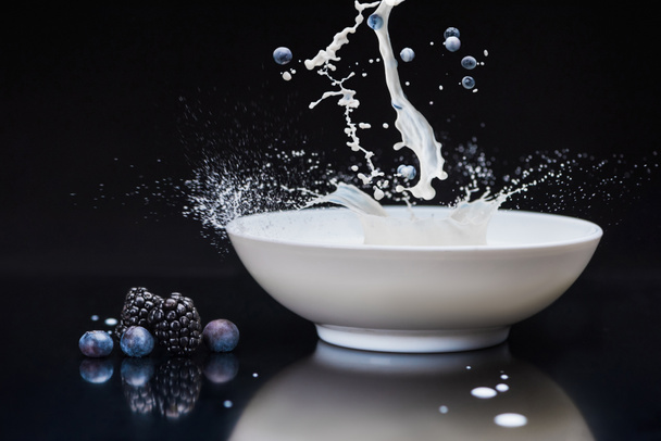 Φρέσκα μούρα εκτινάξεις σε λευκό μπολ με το γάλα σε μαύρο φόντο - Φωτογραφία, εικόνα