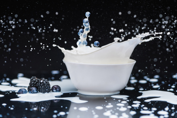 Raw blue berries splashing into white bowl with milk on black background - Foto, afbeelding