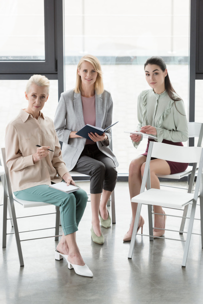 prachtige vrouwelijke ondernemers zitten op stoelen en kijken naar camera in kantoor - Foto, afbeelding