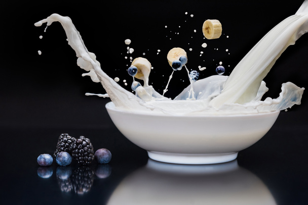 Blackberries and banana dropping into white bowl with milk on black background - Photo, Image