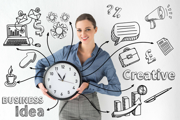 Улыбающаяся деловая женщина, держащая настенные часы в руках на белом с креативной надписью бизнес-идеи и ручными иконами
 - Фото, изображение