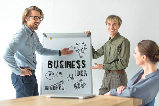 Los hombres de negocios riendo mostrando la inscripción de la idea de negocio y los iconos en flipchat a la mujer de negocios
 - Foto, Imagen