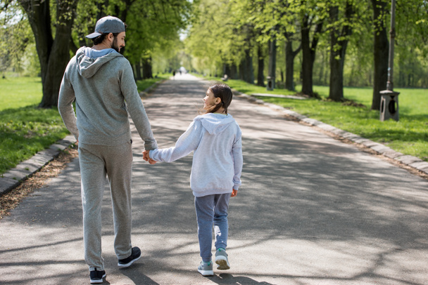 rear view of father and daughter holding hands and walking on path in park  - Фото, изображение
