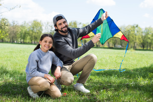 Gelukkig vader en dochter zitten met kite op weide in park - Foto, afbeelding