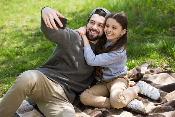 smiling father taking selfie with daughter on plaid in park - Photo, Image