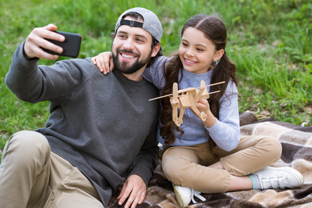 Vater macht Selfie mit Tochter, die Holzspielzeug auf Plaid im Park hält - Foto, Bild