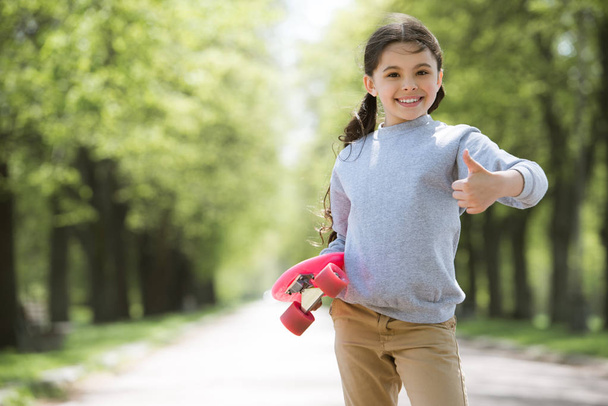 smiling child with skateboard doing thumb up gesture in park - Photo, Image