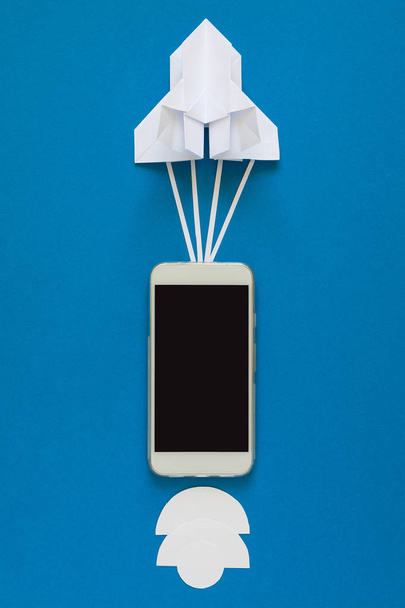 Start up or fast connection concept. The launch paper rocket with smart phone on blue sky with clouds. Origami. Paper cut. Top view - Фото, изображение