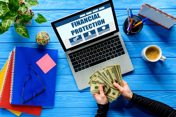 Business person counting dollars by laptop on blue wooden table with stationery, Financial protection lettering - Photo, Image